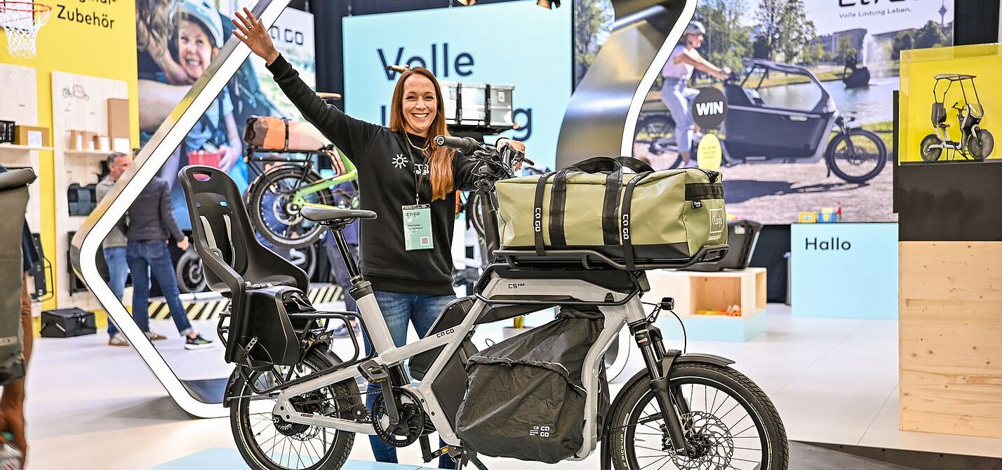 Eine Frau steht an einem Messestand und präsentiert ein Lastenrad. 